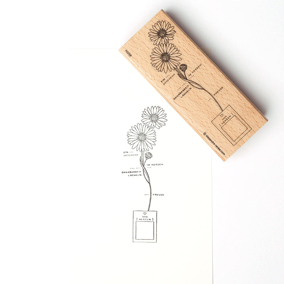 Ein Alexandra Renke Holzstempel und sein Druck zeigen Gänseblümchen, die aus einem Kasten mit deutschem Text auf weißem Papier sprießen - ideal für Papierprojekte und kreative Stempelgummi-Kunstwerke.