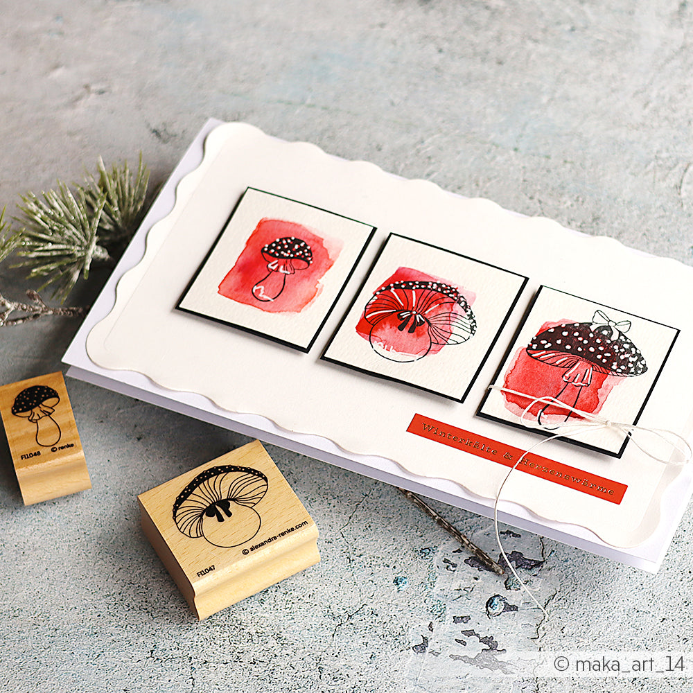 Handgemachte Karte von Alexandra Renke mit drei roten Pilzillustrationen und zwei Holzstempeln auf einem hellen Tisch - perfekt für einzigartige Papierprojekte.