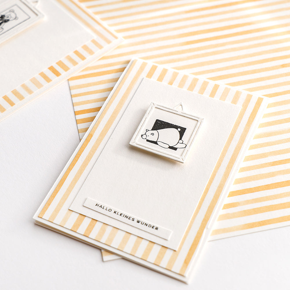 Minimalistische Karte von Alexandra Renke mit einer kleinen Bären-Illustration, Stempelgummi oder Holzstempel-Technik und dem Text "HALLO KLEINES WUNDER" auf einem gestreiften gelben Hintergrund. Ideal für Papierprojekte.
