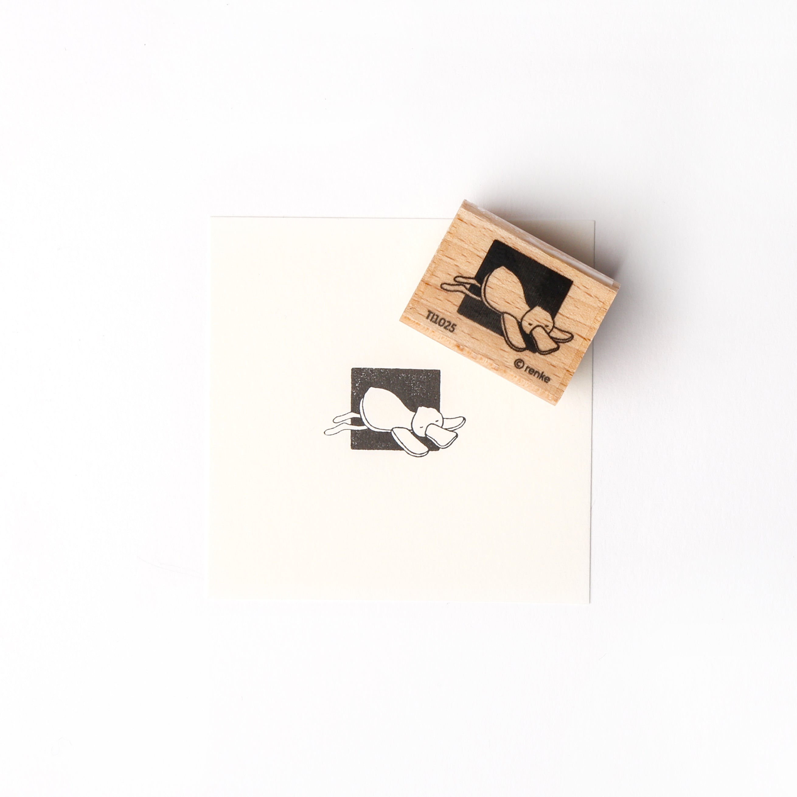 Ein Alexandra Renke Holzstempel mit Stempelgummi stempelt einen schlafenden Entchen-Cartoon vor einem schwarzen Quadrat liegend auf Papier.