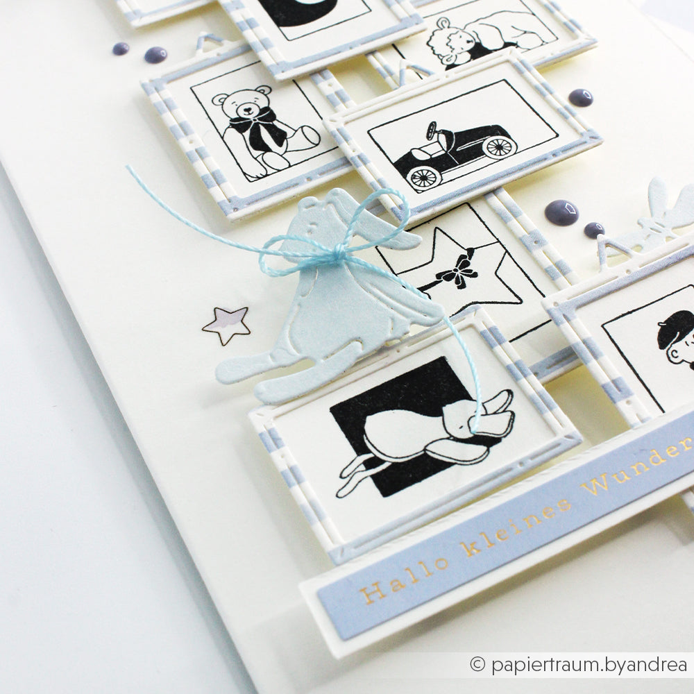 Die handgefertigte Karte von Alexandra Renke zeigt kleine gerahmte Zeichnungen von Spielzeugen und Tieren, eine blaue Schleife, den Text "Hallo kleines Wunder" und ist mit einem Holzstempel und Stempelkissen für zusätzlichen Charme versehen.