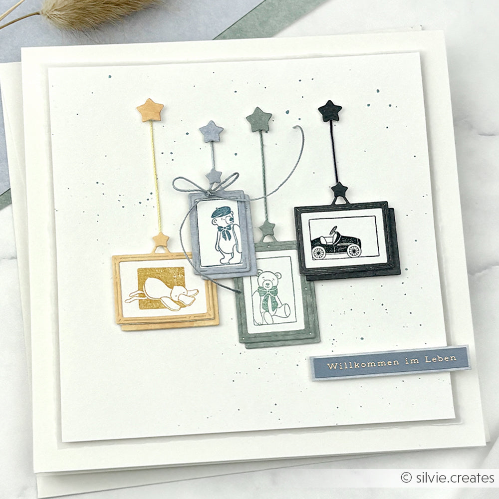 Quadratische Karte von Alexandra Renke mit vier hängenden Rahmen mit Babymotiven, einem deutschen "Willkommen im Leben"-Etikett und zarten Details, die mit Stempelkissen und Holzstempel gestempelt wurden, für ein charmantes Finish.