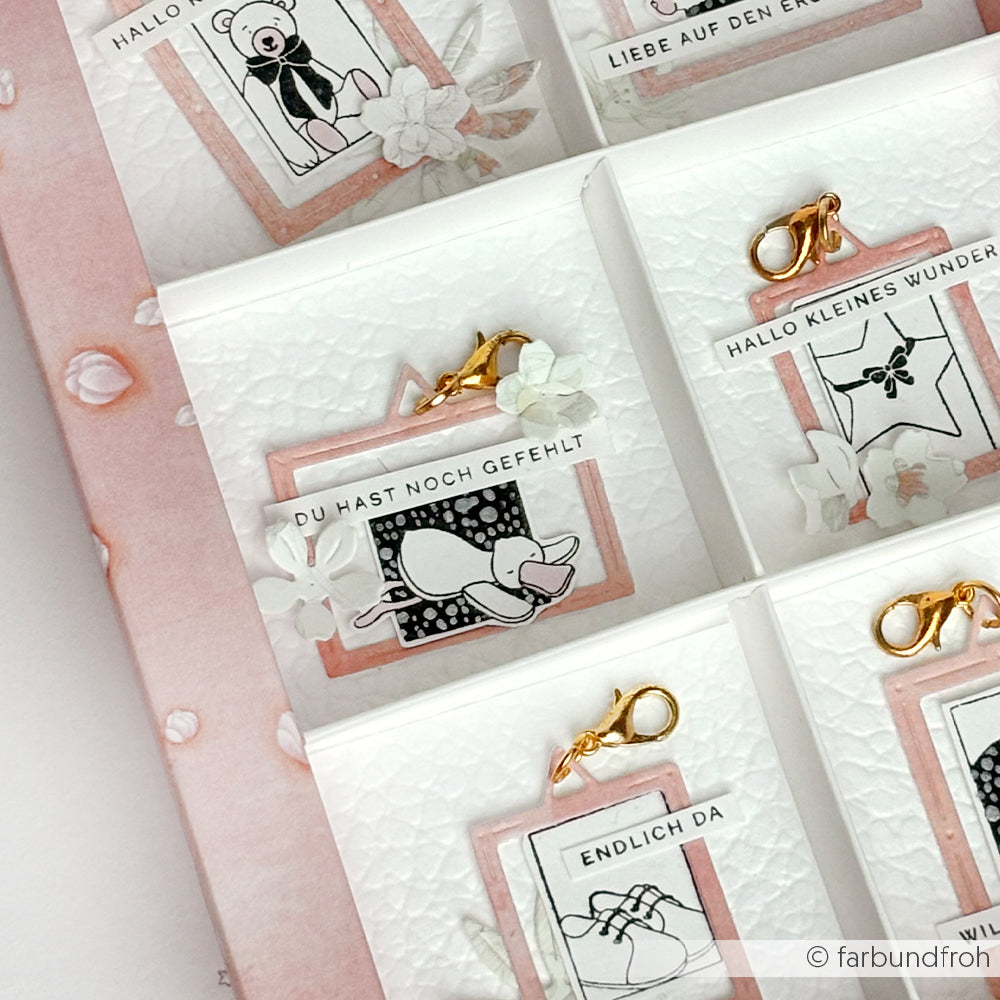 Eine Nahaufnahme der dekorativen Karten von Alexandra Renke mit goldenen Klammern, floralen Details und deutschem Text wie "Schlafendes Entchen" auf rosa-weißem Hintergrund, ideal für die Darstellung von Stempelgummi oder Holzstempel-Designs.