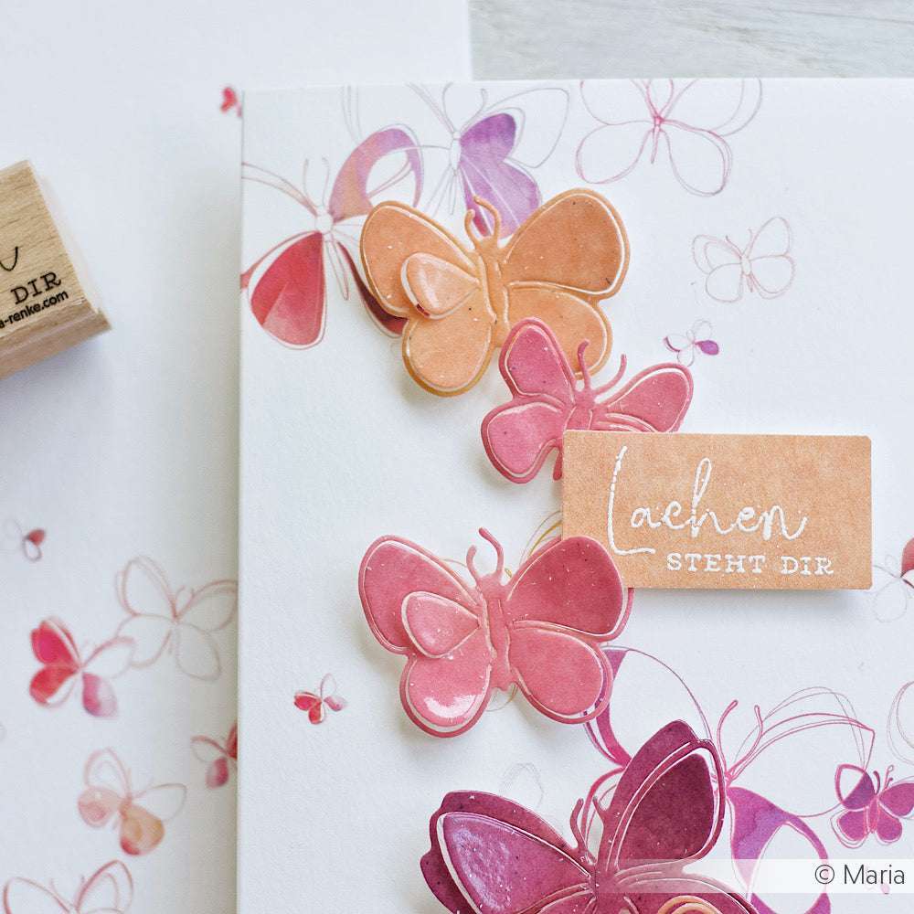 Handgemachte Karte von Alexandra Renke mit rosa und orangefarbenen Schmetterlingen und einem "Lachen steht dir"-Holzstempel - ideal für einzigartige Papierprojekte.