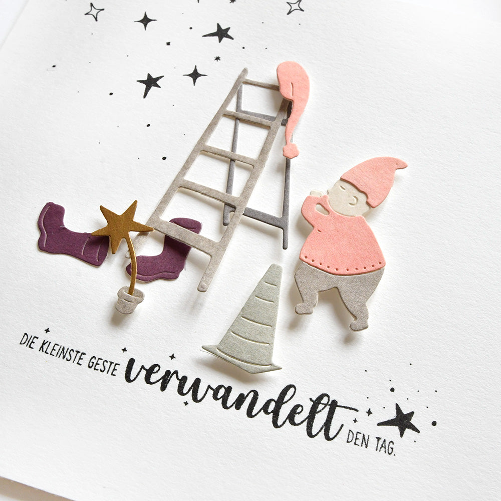 Ein rosafarbener Zwerg bewegt sich auf einer Leiter zu einem Hut und einem Stern, mit deutschem Text, Sternenkritzeleien und Stempelgummi" auf weißem Papier - ein charmanter Entwurf von Alexandra Renke für Papierprojekte.