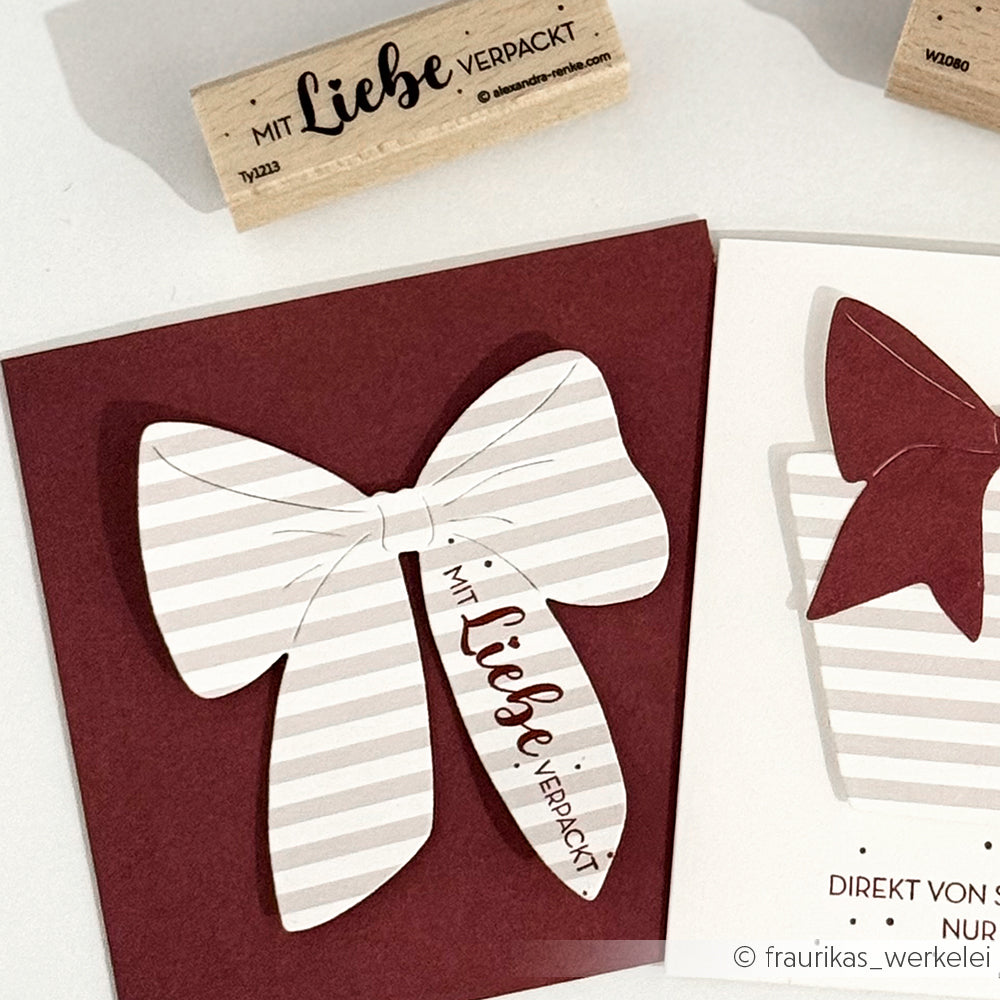 Gestreifte Papierschleifen auf kastanienbraunen und weißen Alexandra Renke Karten, eine Schleife mit der Aufschrift "Mit Liebe verpackt". Hochwertiges Holz, Holzstempel und Stempelkissen sind in der Nähe für kreative Geschenkverpackungen angeordnet.