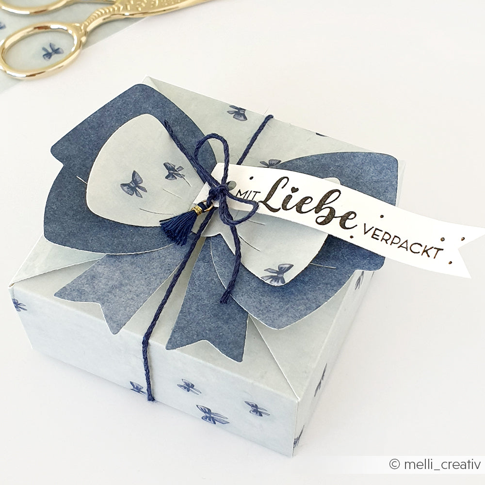 Eine blau-weiße Geschenkschachtel mit Schleife, Schnur und "Mit Liebe verpackt"-Anhänger von Alexandra Renke - perfekt, um sie mit ihrem Stempelgummi oder Holzstempel zu kombinieren und ihr eine persönliche Note zu verleihen.