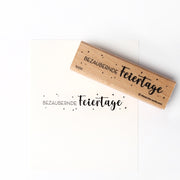 Alexandra Renke Holzstempel und Papier mit dem Schriftzug "Bezaubernde Feiertage" in deutscher Sprache, umgeben von Sternen - ein detailliertes Design, das perfekt ist, um eine charmante festliche Note zu verleihen.