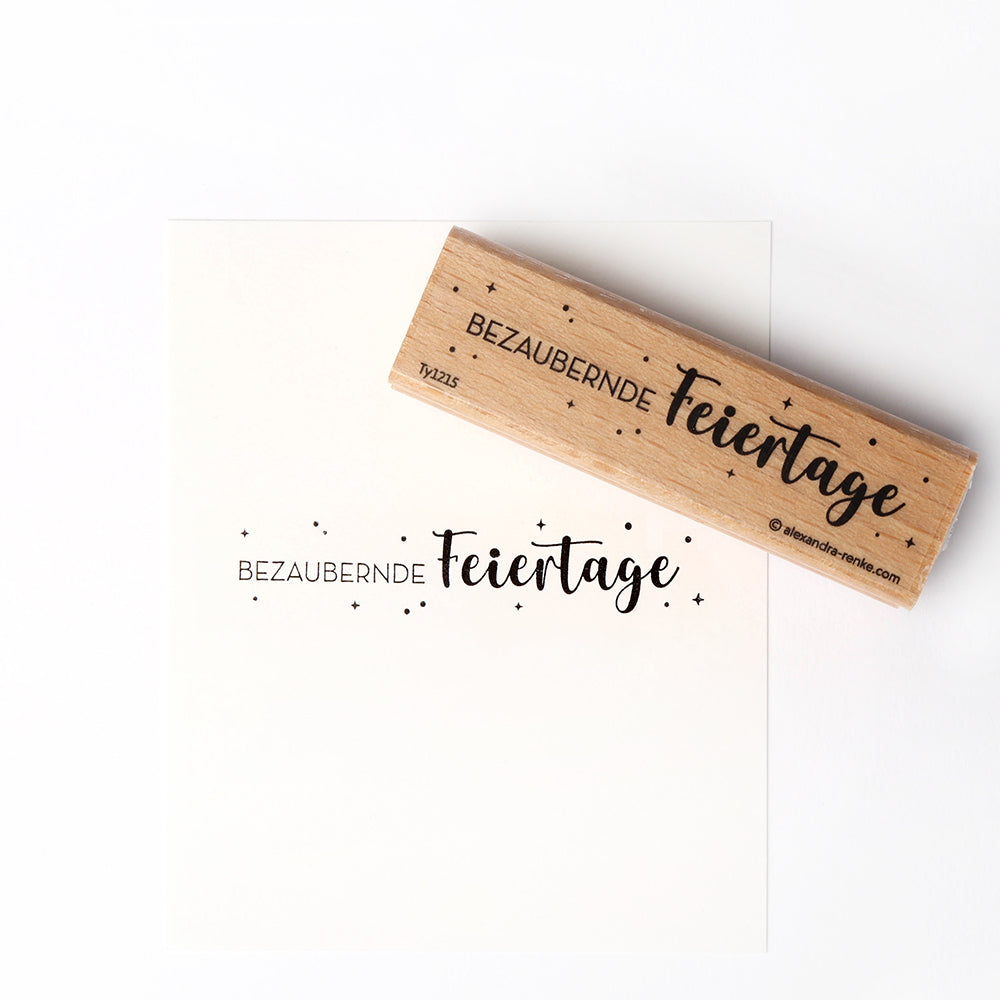 Alexandra Renke Holzstempel und Papier mit dem Schriftzug "Bezaubernde Feiertage" in deutscher Sprache, umgeben von Sternen - ein detailliertes Design, das perfekt ist, um eine charmante festliche Note zu verleihen.