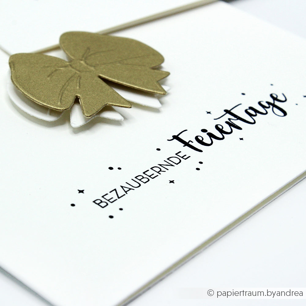 Weiße Karte von Alexandra Renke mit einer goldenen Schleife und dem Text "Bezaubernde Feiertage" in Schwarz, mit detaillierten Motiven aus einem hochwertigen Holzstempel.