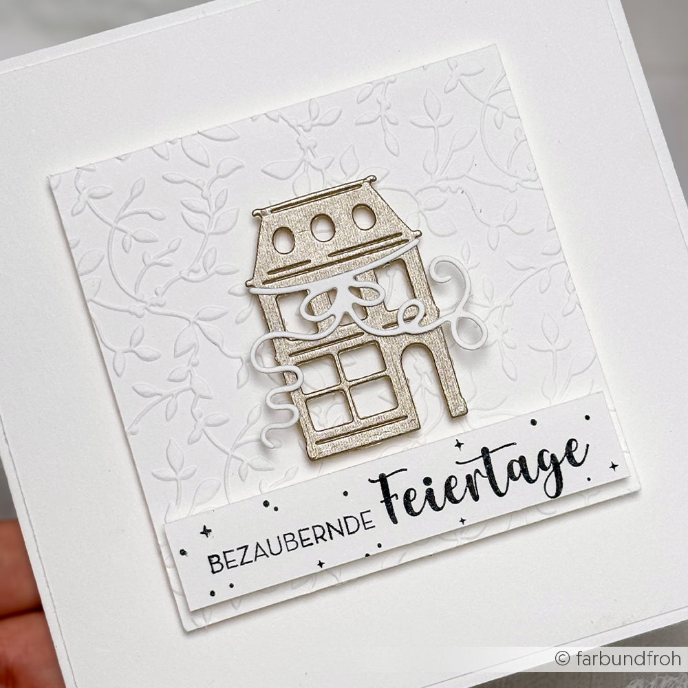 Weiße Grußkarte von Alexandra Renke mit geprägtem Blatthintergrund und ausgestanztem Haus über dem Stempelgummi-Text "Bezaubernde Feiertage".