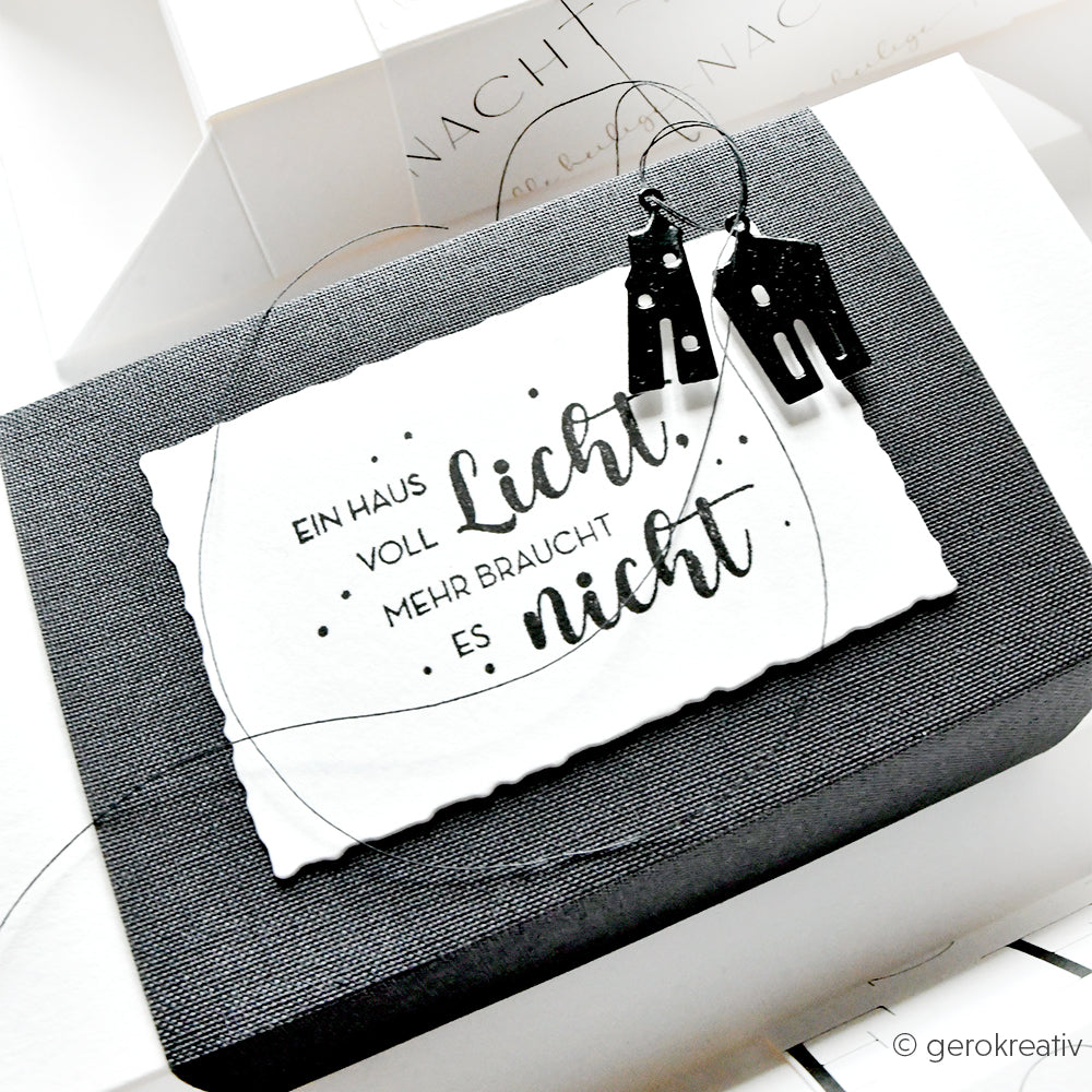 Schwarz-weiße Karte mit deutschem Text und schwarzen Ohrringen in Form eines Hauses auf einer strukturierten Schachtel, mit Alexandra Renke Holzstempel mit detailreichen Motiven für zusätzlichen Charme.