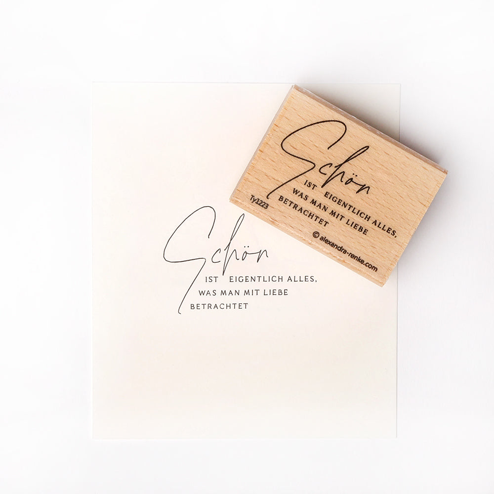 Ein Holzstempel von Alexandra Renke und gestempeltes Papier mit deutschem Text in eleganter Schrift auf weißem Hintergrund.