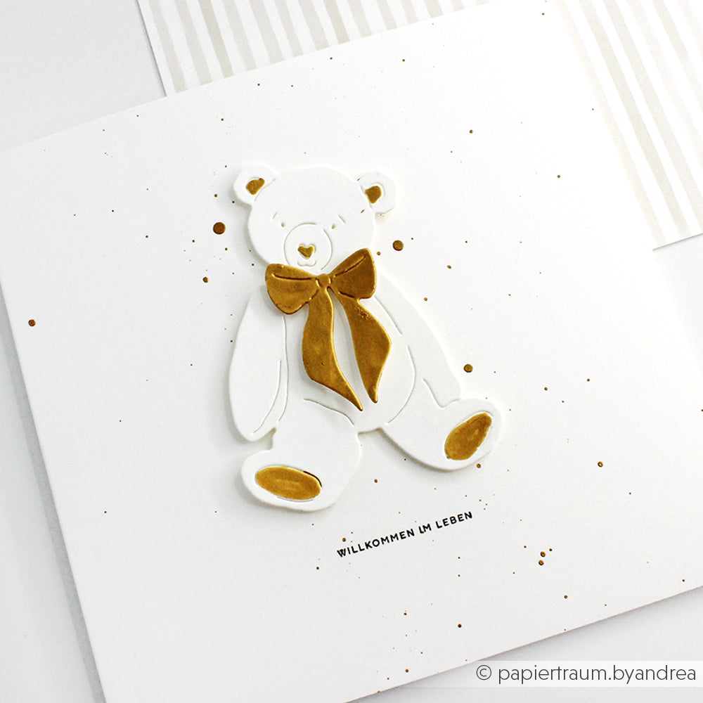Weiße Karte von Alexandra Renke mit einem goldfarbenen Teddybär, gestempelt mit Stempelgummi oder Holzstempel, und dem deutschen Gruß "Willkommen im Leben".