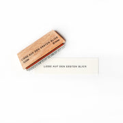 Ein Holzstempel und Papier von Alexandra Renke zeigen den deutschen Satz "LIEBE AUF DEN ERSTEN BLICK" auf weißem Hintergrund, perfekt für Papierprojekte.