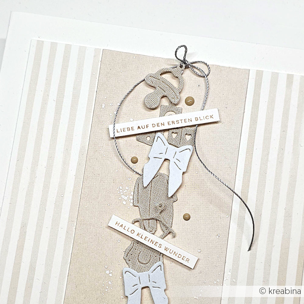 Eine handgefertigte Karte von Alexandra Renke mit Schleifen, einem Schnuller und deutschem Text feiert ein Neugeborenes auf einem beigen Hintergrund.
