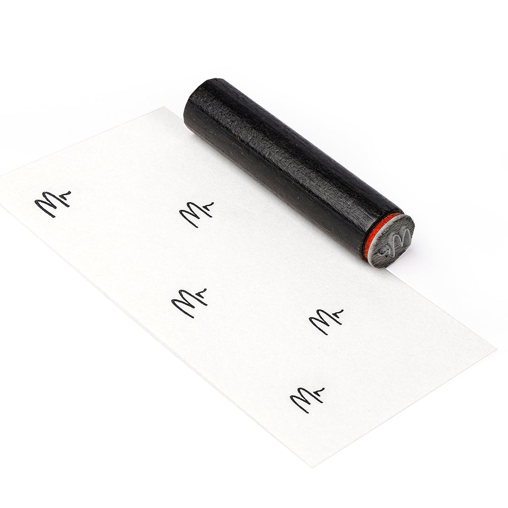 Ein Alexandra Renke Holzstempel mit einem M-Motiv stempelt den Buchstaben M wiederholt auf ein weißes Blatt Papier.