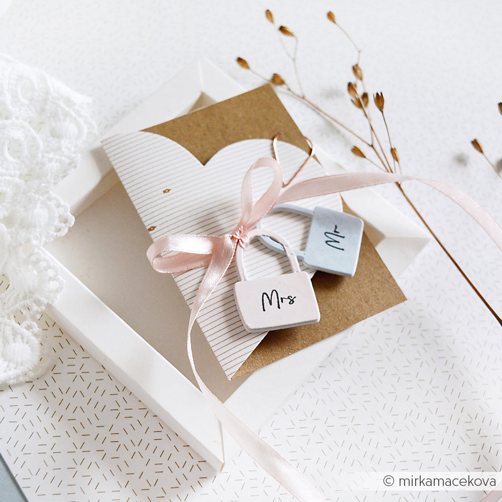 Eine Hochzeitskarte von Alexandra Renke mit "Mr"- und "Mrs"-Anhängern, rosa Schleife, einer weißen Schachtel, Trockenblumen und einem eleganten Akzent-Stempel-Motiv auf einer gemusterten Oberfläche.