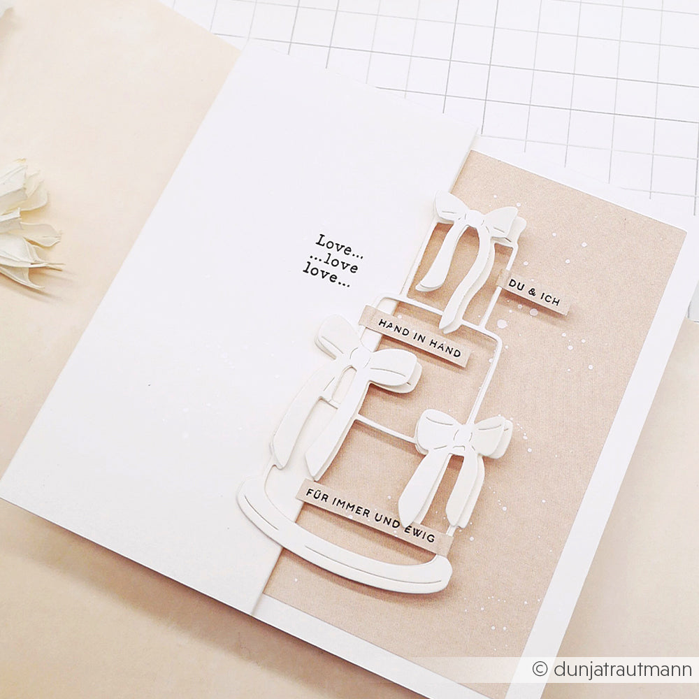 Minimalistische Hochzeitskarte von Alexandra Renke mit weißer Torte, Schleifen und deutschen Sprüchen auf neutralem Hintergrund, mit zarten Mini Typo Hand in Hand Details und charmanten Stempel Holz Akzenten.