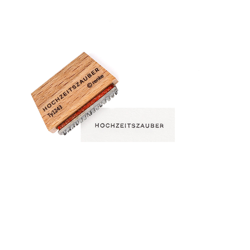 Ein Holzstempel von Alexandra Renke mit Stempelgummi und der Aufschrift HOCHZEITSZAUBER liegt neben einem Papier, das ebenfalls mit Hochzeitszauber bestempelt wurde.