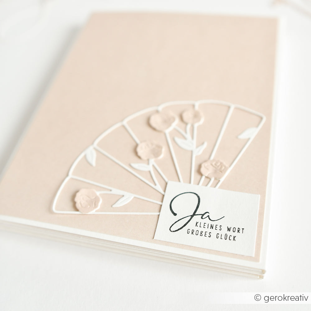 Die beigefarbene Hochzeitskarte von Alexandra Renke zeigt ein weißes Blumenfächermotiv, ist mit einem Holzstempel und Stempelkissen bestempelt und enthält ein Etikett "Ja kleines Wort großes Glück".