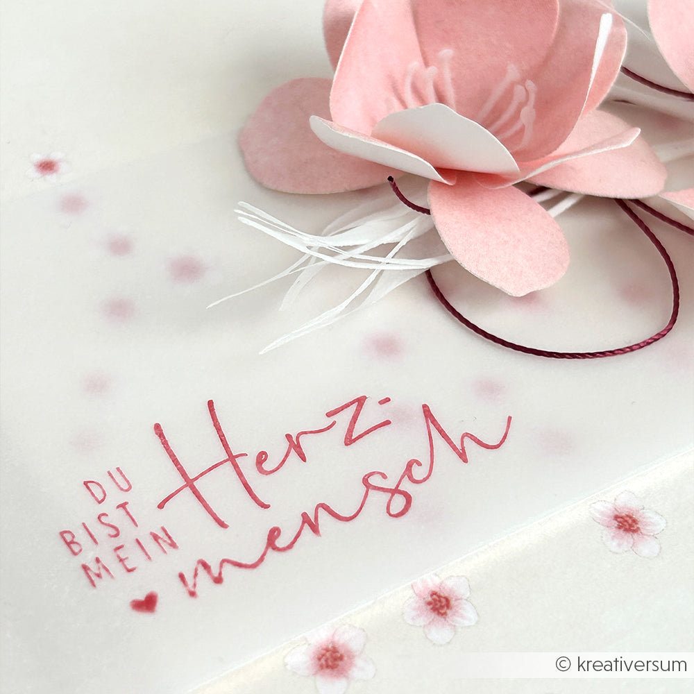 Eine rosafarbene Papierblume, gestempelt mit "Du bist mein Herzensmensch" mit einem Holzstempel von Alexandra Renke, vor einem weißen, blumengemusterten Hintergrund.
