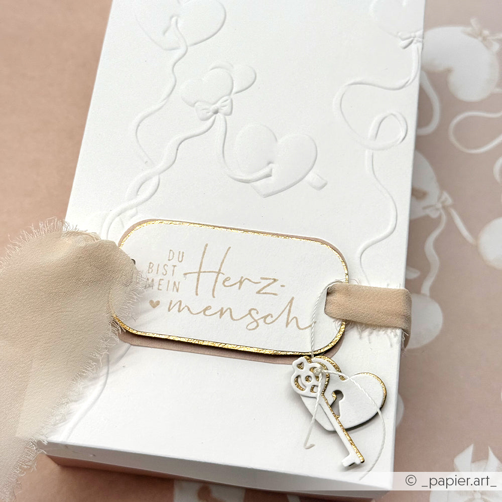 Weiße Karte von Alexandra Renke mit geprägten Herzluftballons, beigem Band, Schlüsselanhänger und deutschem Text "Du bist mein Herzmensch". Mit detailliertem Design und passendem Holzstempel für einen besonderen Herzmensch-Moment.