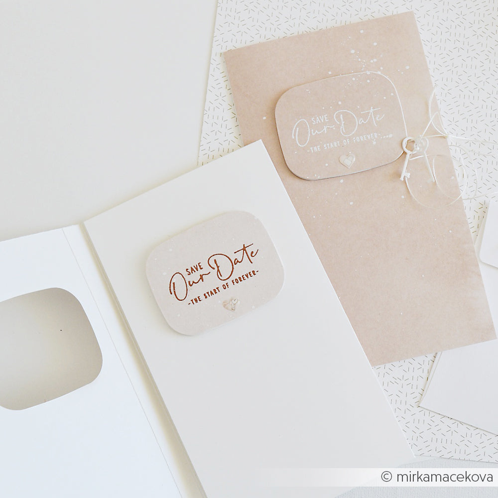 Zwei elegante Save Our Date-Karten von Alexandra Renke, eine weiße und eine beige, werden auf einer weißen Fläche neben einem klassischen Holzstempel präsentiert.
