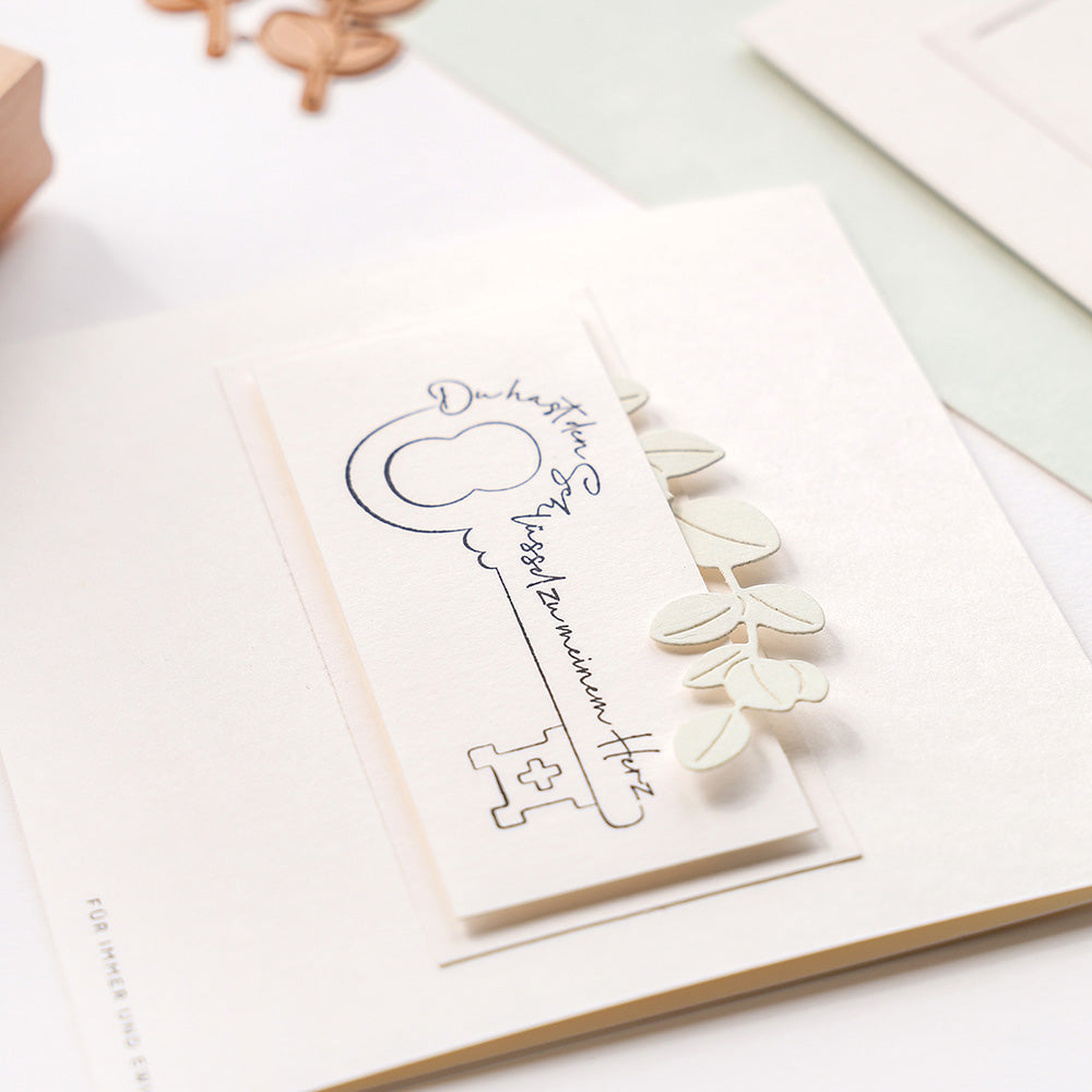 Eine handgefertigte Karte von Alexandra Renke mit einem handgezeichneten Schlüssel, elegantem kursivem Text und kleinen Papierblättern auf weißem Grund, mit charmanten Akzenten durch Stempelgummi und Stempelkissen.