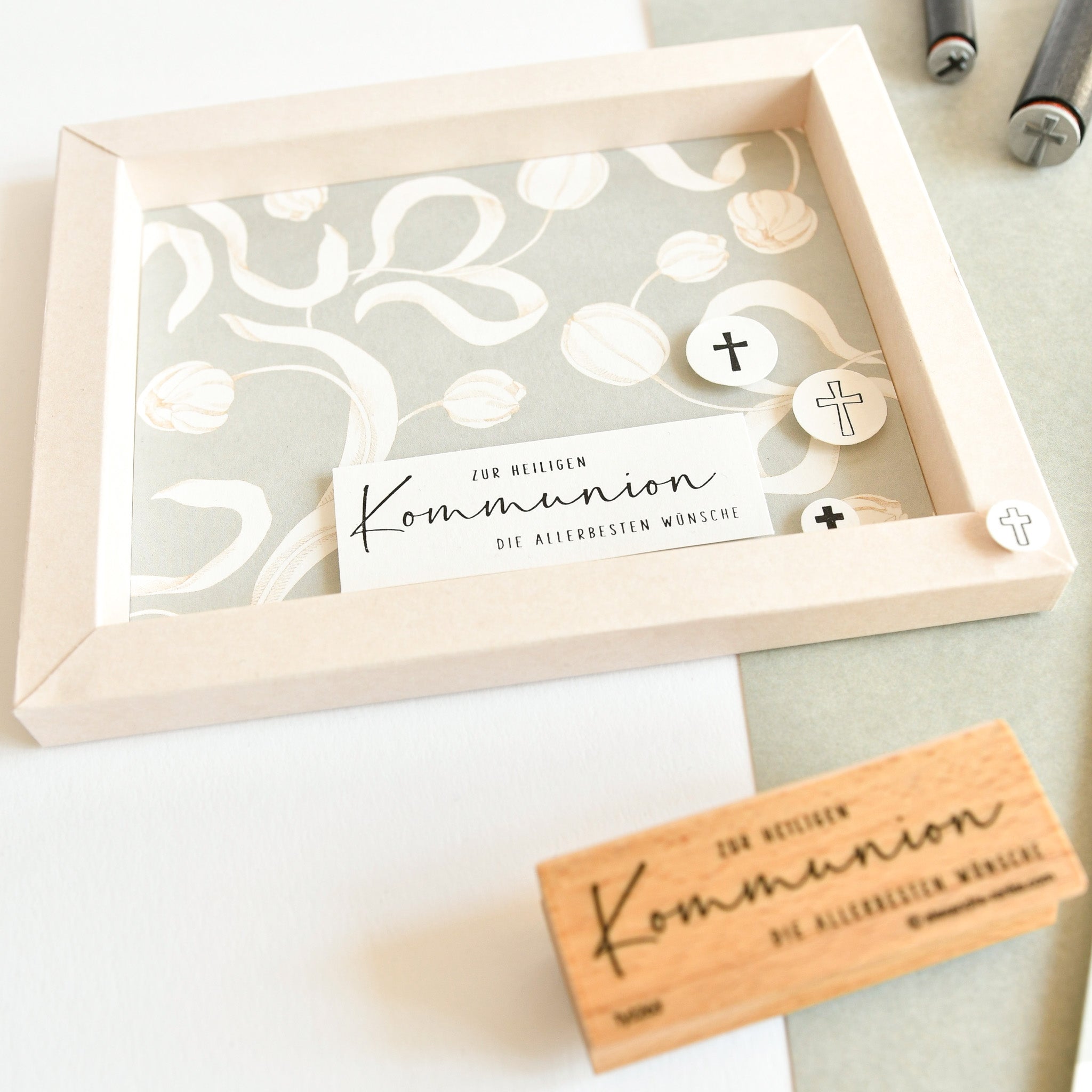 Gerahmte Karte von Alexandra Renke mit dem Text "heilige Kommunion", einem dekorativen Hintergrund und einem Holzstempel, die auf einer hellen Fläche angebracht ist.