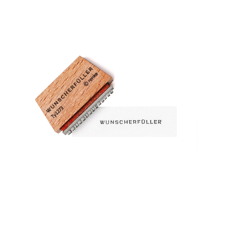 Ein Holzstempel von Alexandra Renke mit der Aufschrift WUNSCHERFÜLLER stempelt das Wort auf weißes Papier und hebt die feinen Details des Stempelgummis hervor.