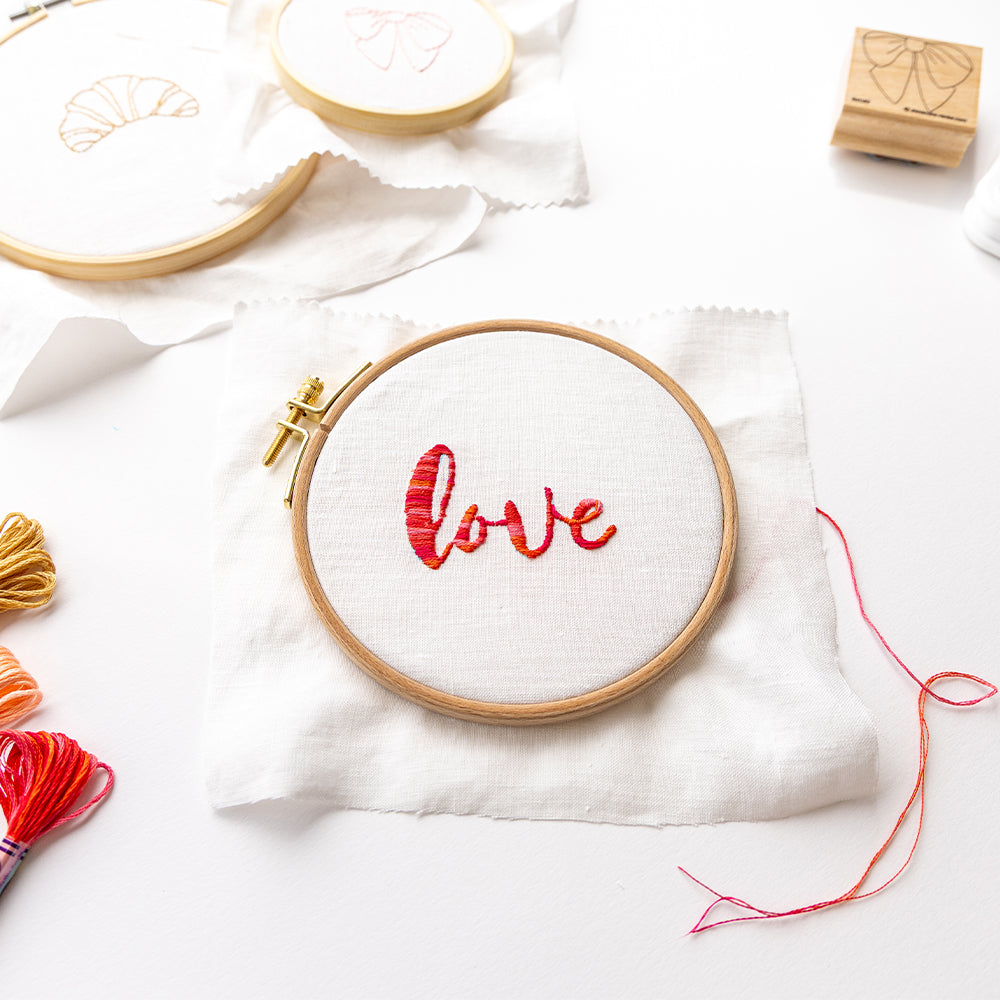 Alexandra Renke Stickrahmen mit dem Motiv "Liebe" in Rot auf weißem Stoff, umgeben von Vorräten und einem Stickvorlagen Stempel für mühelose Musterübertragung.