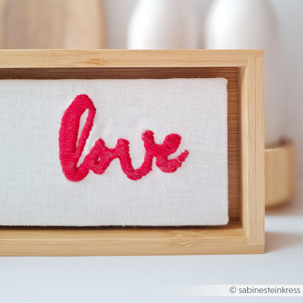 Weißer Stoff mit rot gesticktem "love", gerahmt in einer hellen Holzbox von Alexandra Renke - eine ideale Inspiration für individuelle Stickprojekte oder als Sticken einfacher Anleitung.