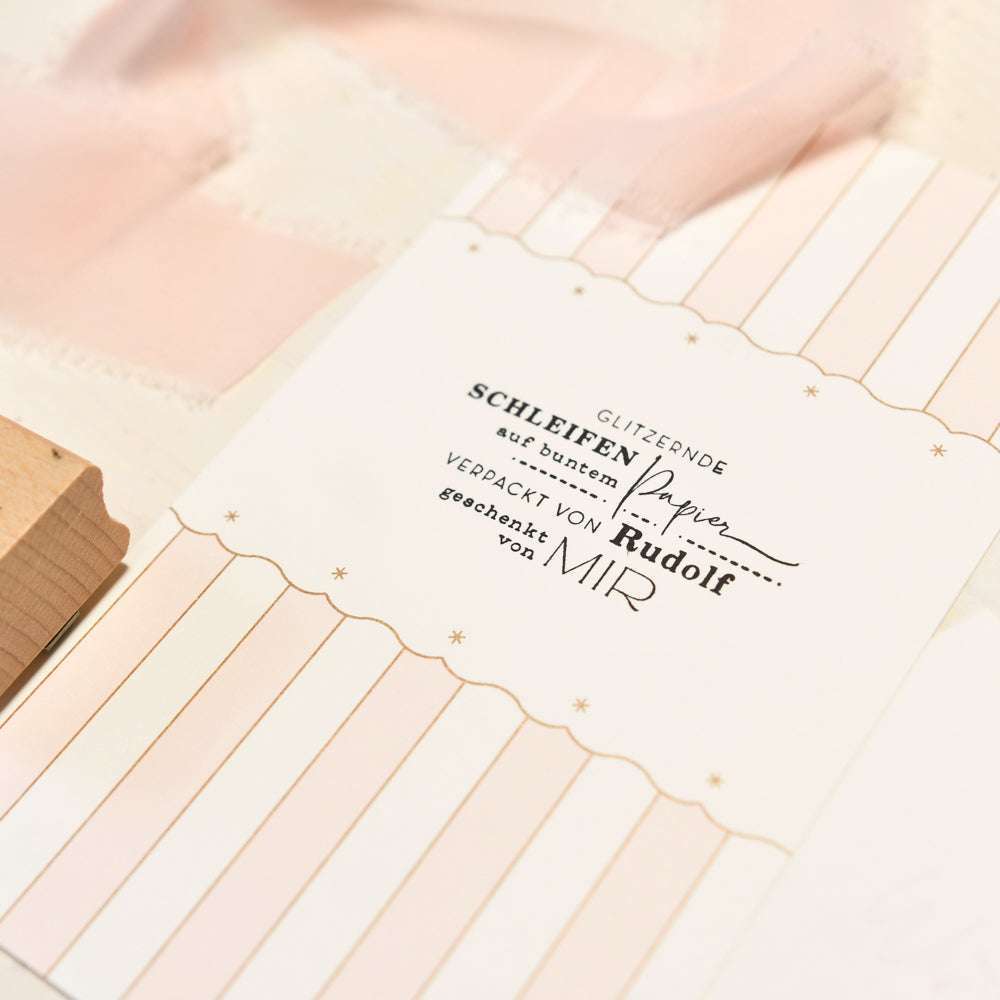 Eine gestreifte Karte von Alexandra Renke mit deutschem Text, einer rosa Schleife und einem Holzstempel auf einer hellen Oberfläche.