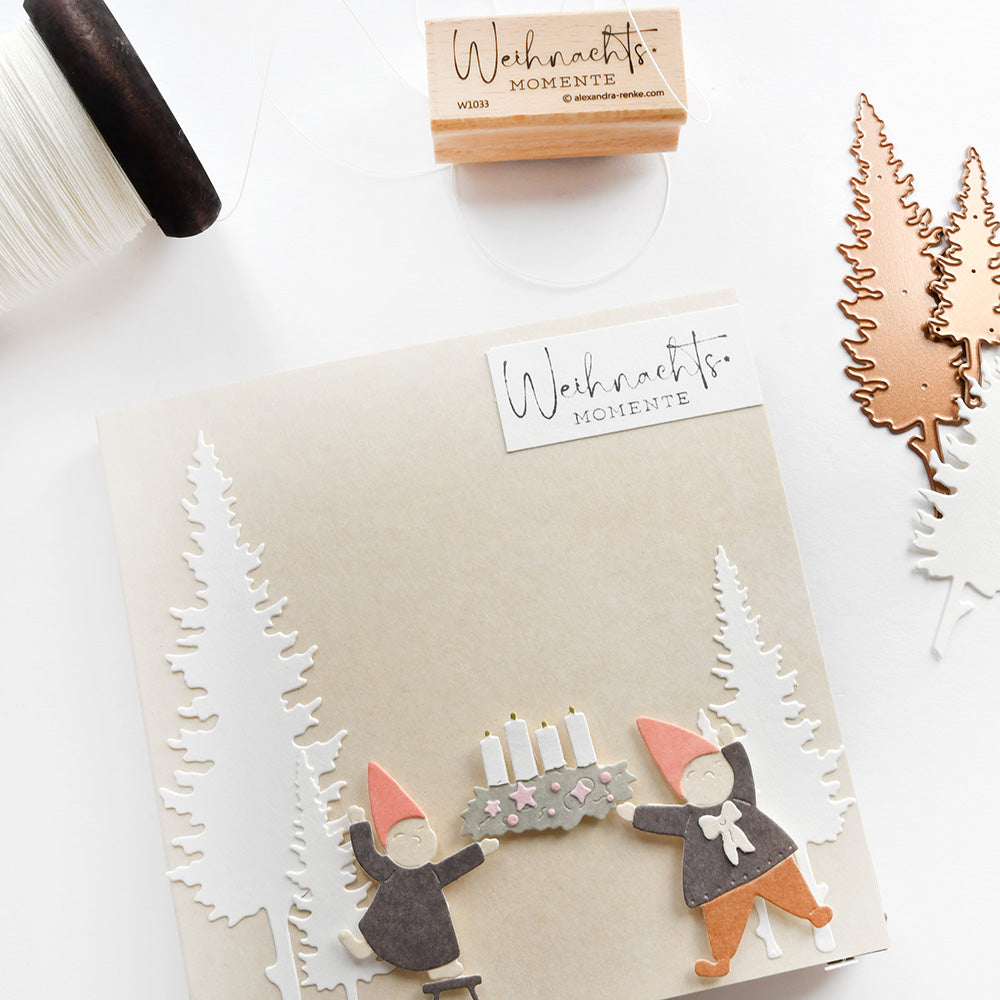 Handgefertigte Weihnachtskarte von Alexandra Renke mit zwei Wichteln, einem Kerzenkranz, festlichen Bäumen, einem herzlichen "Weihnachtsmomente"-Text und schönen Stempelgummi-Akzenten.