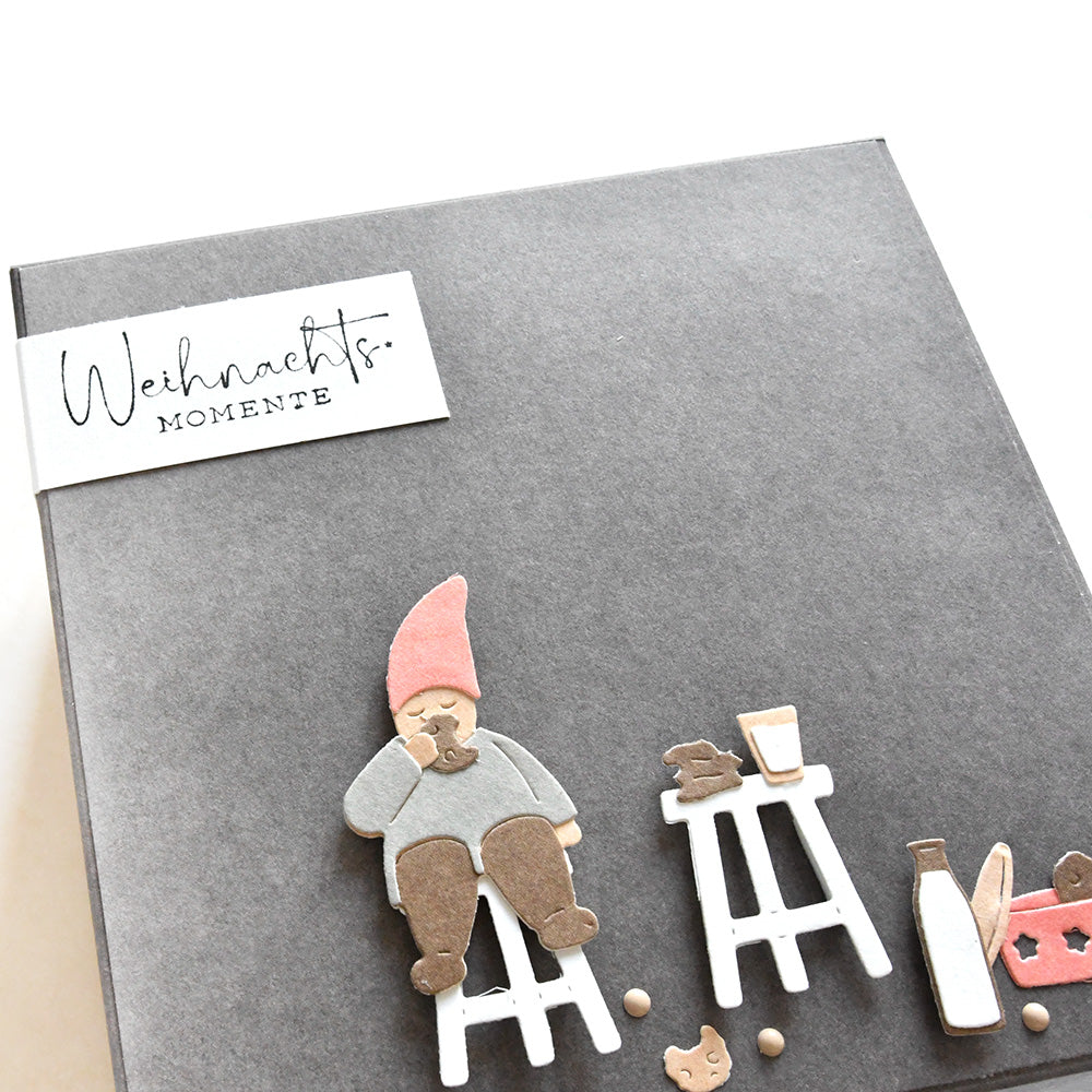 Graue Alexandra Renke Karte mit einem Wichtel auf einem Hocker, Getränken, Leckereien und dem Text "Weihnachtsmomente" - ideal zum Basteln mit Ihrem Lieblings-Stempelgummi oder Holzstempel.
