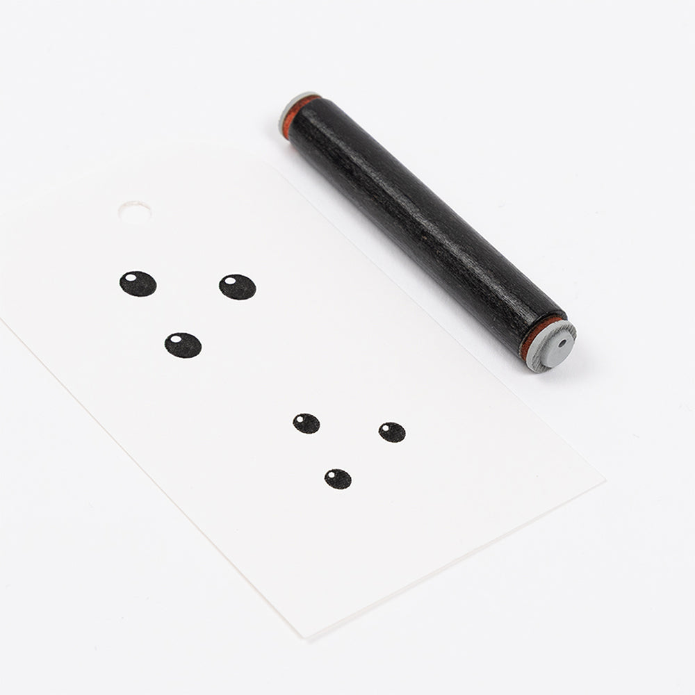 Ein kleines Mini-Beeren-Roller-Werkzeug aus dunklem Holz neben einer weißen Karte mit schwarzen Punkten.