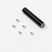 Ein schwarzer, zylindrischer Holzstempel neben einem Etikett, auf dem fünf kleine Stiftsymbole aufgedruckt sind.