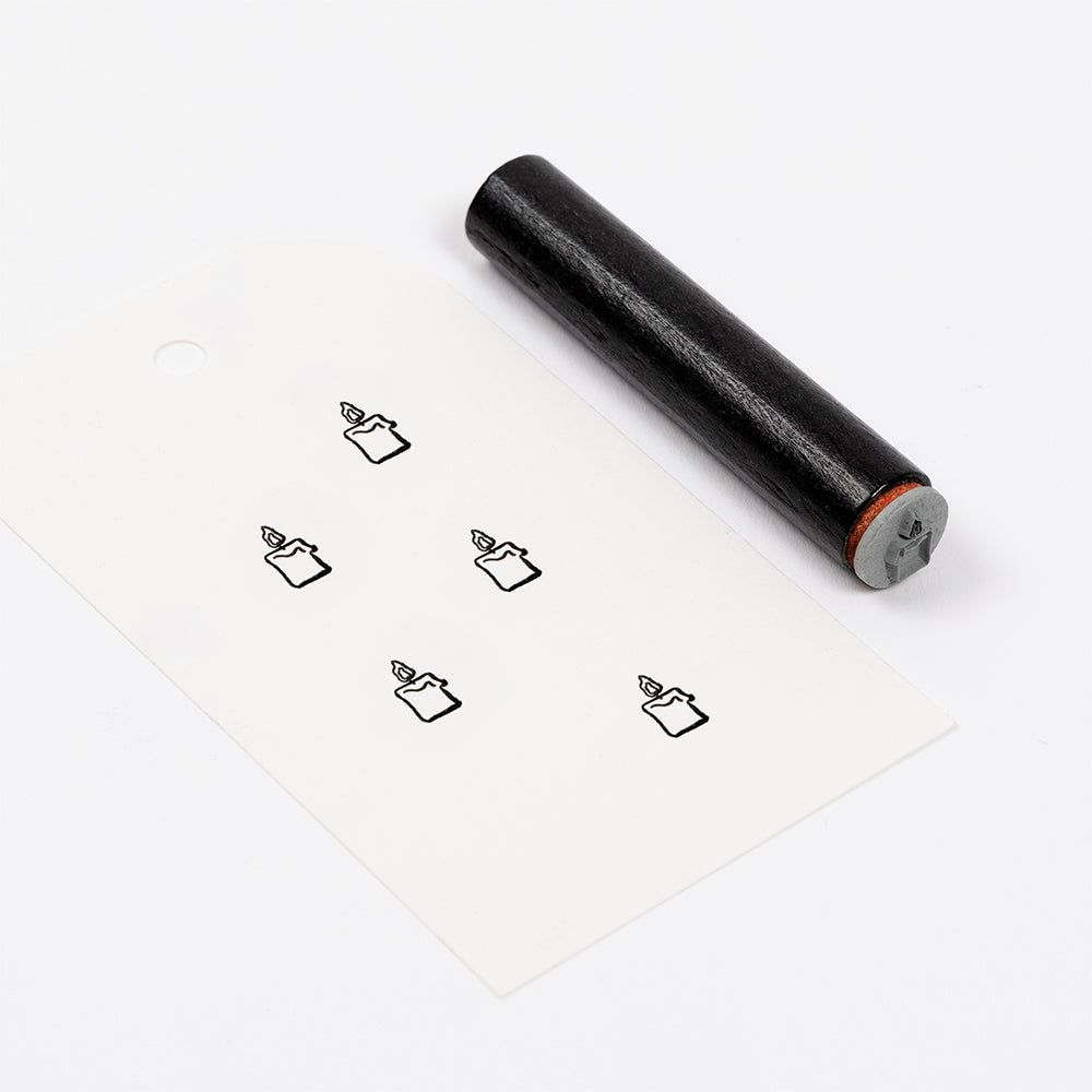 Ein schwarzer, zylindrischer Holzstempel neben einem Etikett, auf dem fünf kleine Stiftsymbole aufgedruckt sind.