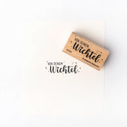 Ein Holzstempel und Papier von Alexandra Renke mit dem deutschen Text "Von deinem Wichtel" in schwarzer Schrift - liebevoll aus hochwertigem Holz gestaltet mit charmantem Stempelmotiv.