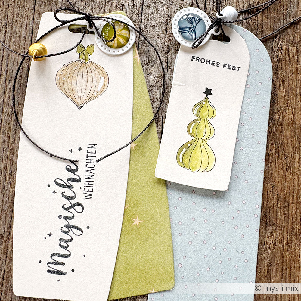 Zwei festliche Weihnachtsgeschenkanhänger von Alexandra Renke mit Ornamenten, dem Text "Magische Weihnachten" und einer Schnur im Holzstempel-Design auf einer Holzfläche.