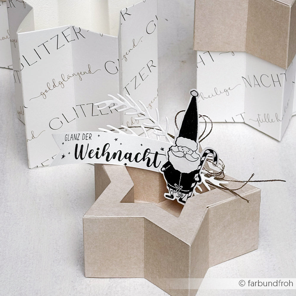 Ein sternförmiger Geschenkkarton von Alexandra Renke mit einem festlichen Anhänger, einem Cartoon-Weihnachtsmann und dem deutschen Text "Glanz der Weihnacht", der neben weißen Kartons angebracht ist.
