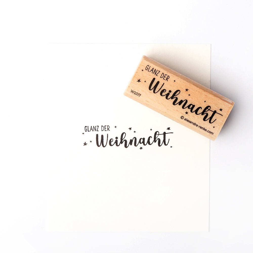 Ein hölzerner Weihnachtsstempel von Alexandra Renke und Papier zeigen beide GLANZ DER Weihnacht mit Sternen in schwarzer Schrift.