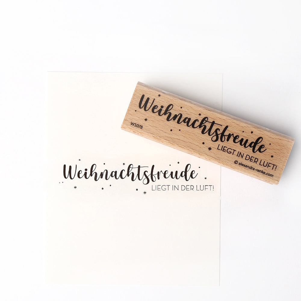 Ein Holzstempel und ein Stempelkissen von Alexandra Renke hinterlassen den Schriftzug "Weihnachtsfreude LIEGT IN DER LUFT!" auf weißem Papier.
