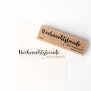 Ein Holzstempel und ein Stempelkissen von Alexandra Renke hinterlassen den Schriftzug "Weihnachtsfreude LIEGT IN DER LUFT!" auf weißem Papier.