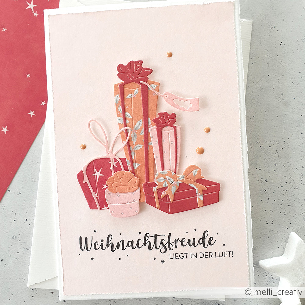 Rosa Karte von Alexandra Renke mit geschichteten Papiergeschenkboxen, einem Cupcake und dem deutschen Text "Weihnachtsfreude liegt in der Luft!" - verziert mit Holzstempeln und Stempelkissen für einen festlichen, handgemachten Look.