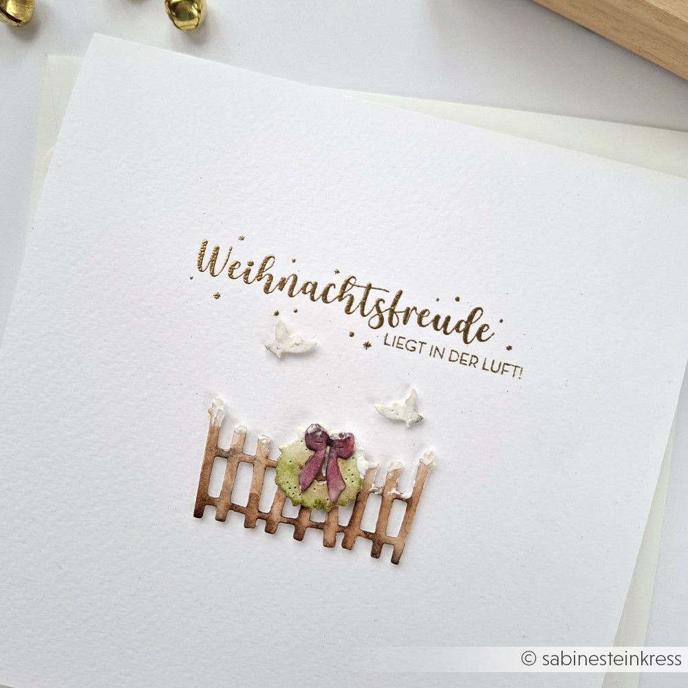 Weiße Karte mit goldenem Text, Holzzaun, Kranz mit Schleife und zwei weißen Vögeln - perfekt, um Weihnachtsfreude zu verbreiten. Mit aufwendigen Details, die an die Handwerkskunst von Holzstempeln erinnern. Von Alexandra Renke.