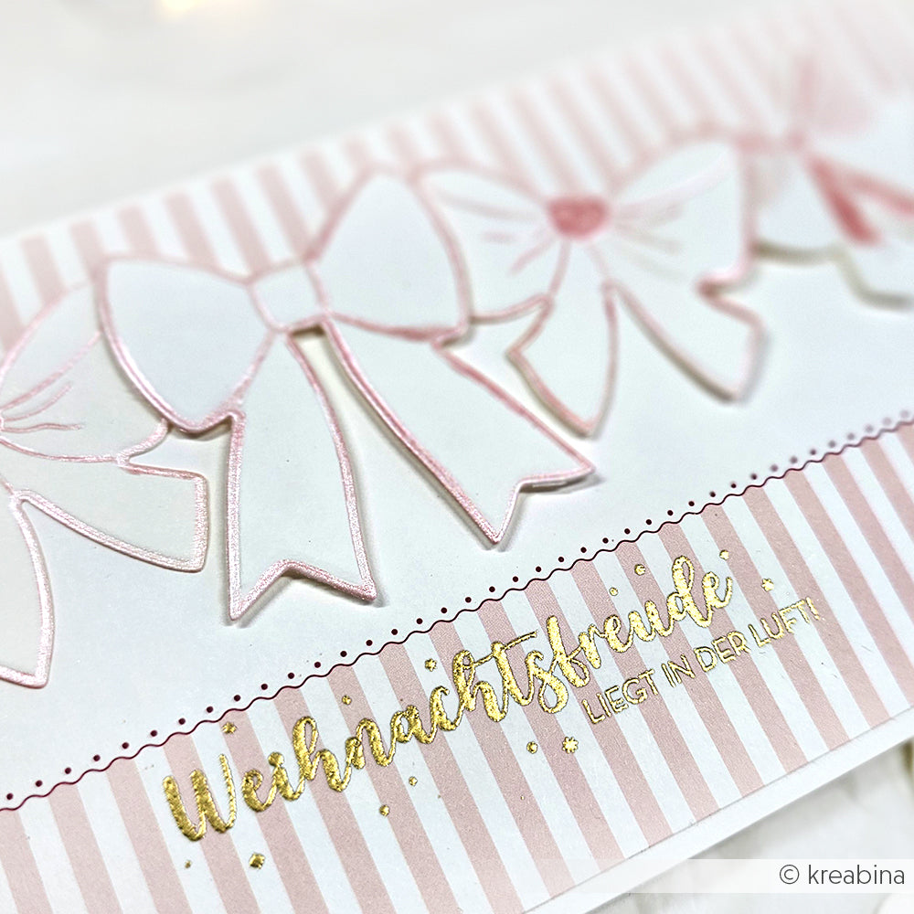 Rosa-weiße Karte mit Schleifen, goldener Text "Weihnachtsfreude liegt in der Luft!" auf Streifen - ideal zum Kombinieren mit Alexandra Renke Holzstempel oder Stempelgummi für festliche Bastelarbeiten.