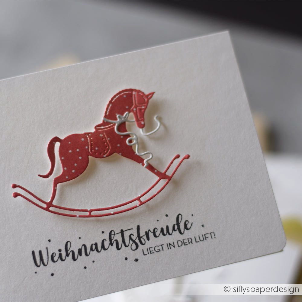 Alexandra Renke weiße Karte mit rotem Schaukelpferd, gestempelt mit "Weihnachtsfreude liegt in der Luft!.