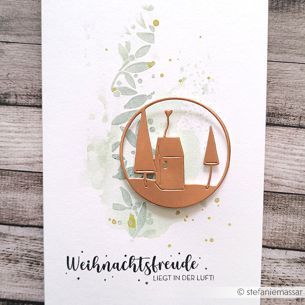 Ein Kupferhaus und Bäume sind auf einem aquarellierten Hintergrund mit dem deutschen Text "Weihnachtsfreude liegt in der Luft!" von Alexandra Renke gestanzt. Perfekt zum Basteln mit Holzstempeln oder Stempelgummi.
